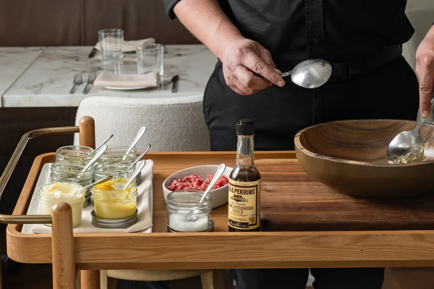 A server mixing steak tartare in a wooden bowl on a tableside cart at L'Étoile in Oyster Bay, with jars of condiments and a bowl of chopped beef