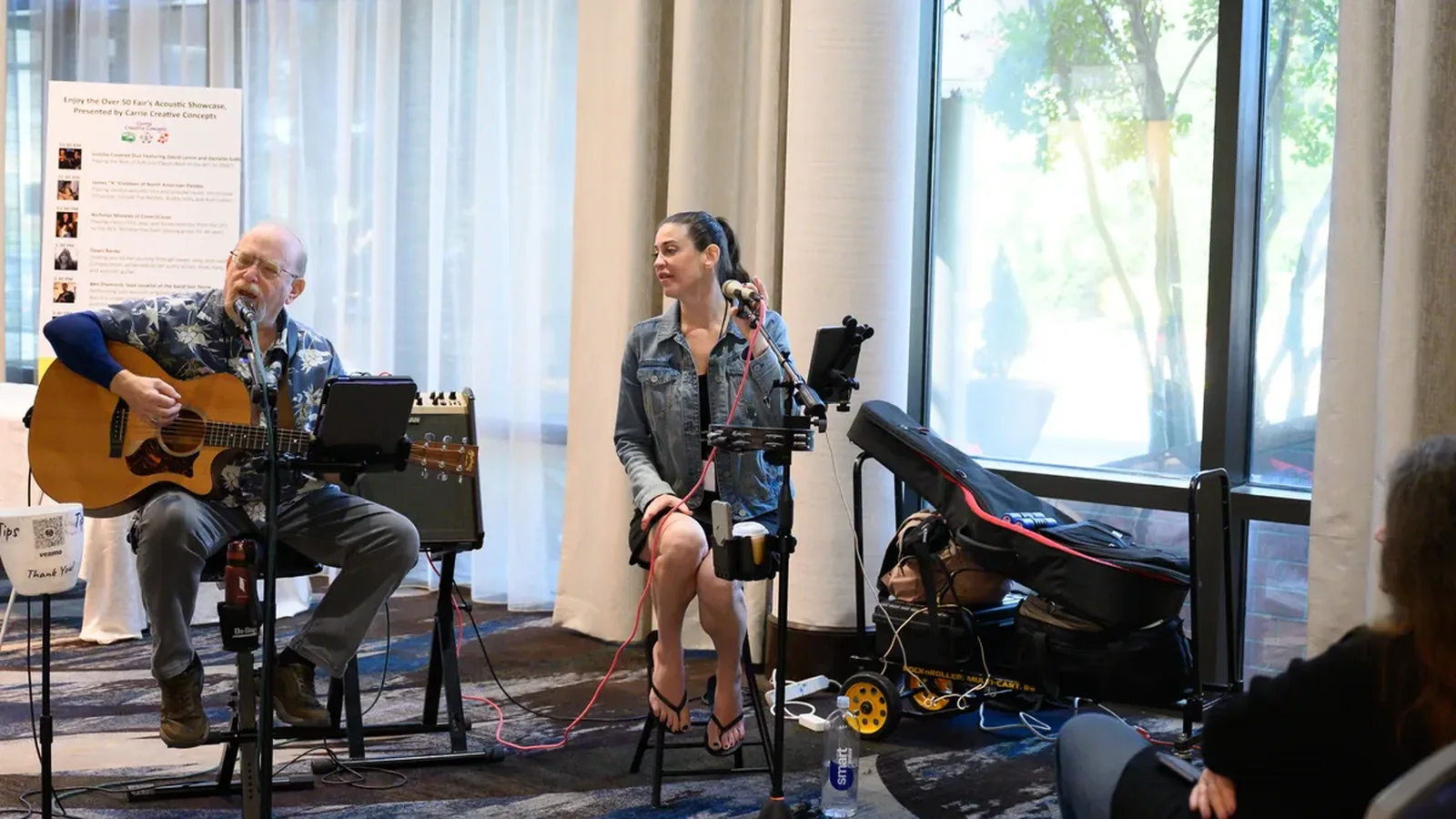 Two musicians performing at the Over 50 Fair, a man playing acoustic guitar and a woman singing into a microphone, beside a window full of daylight