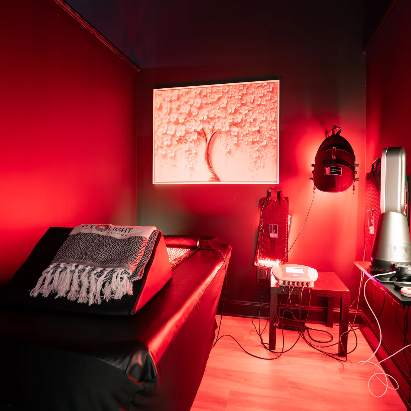 A red light therapy room at Red Light Method Commack, bathed in deep red light, with a treatment bed, a wall-mounted light array and a framed cherry-blossom lightbox