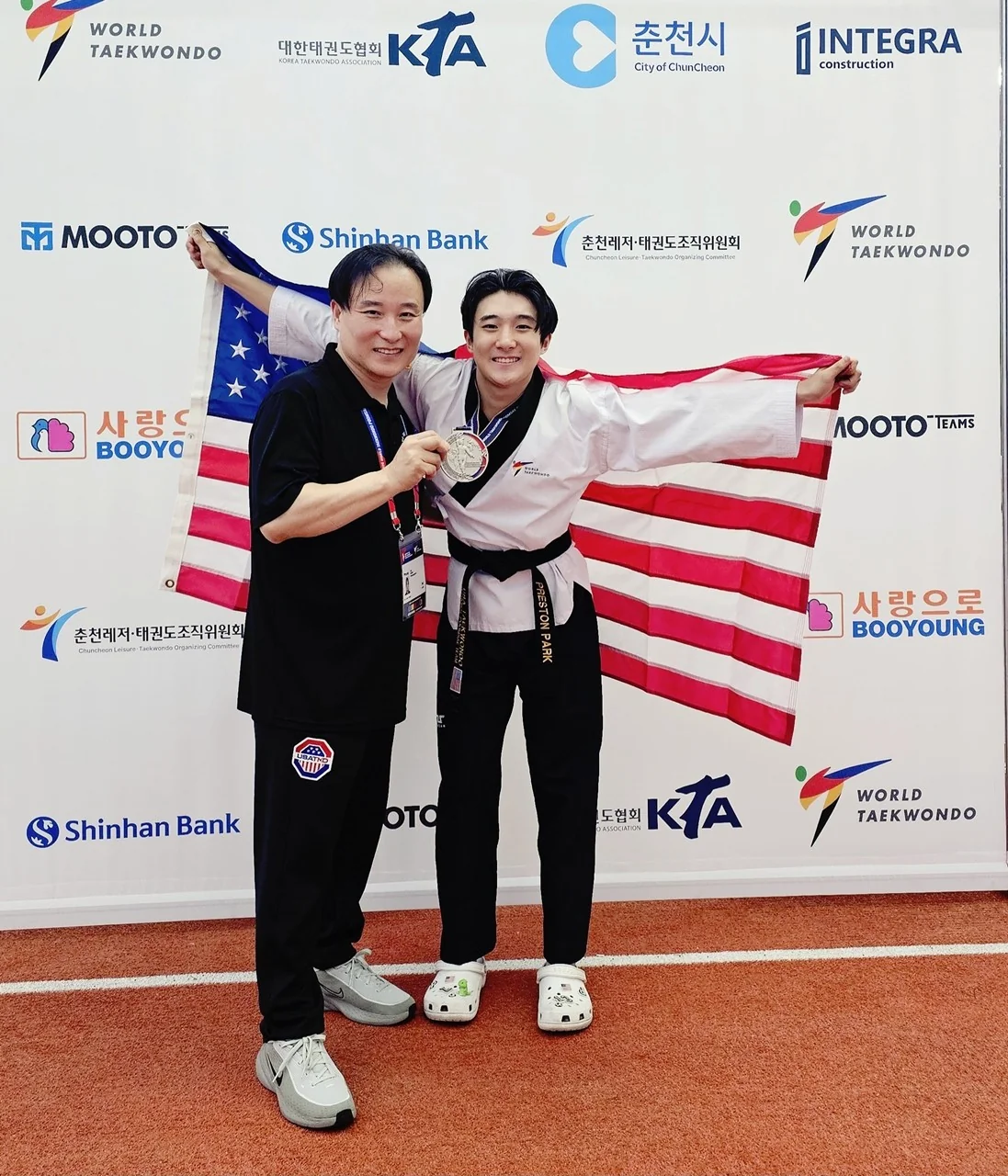 Preston Park in his taekwondo uniform holds an American flag behind him while his father and coach, Master Jay Park, holds up his silver medal in front of a sponsor wall.