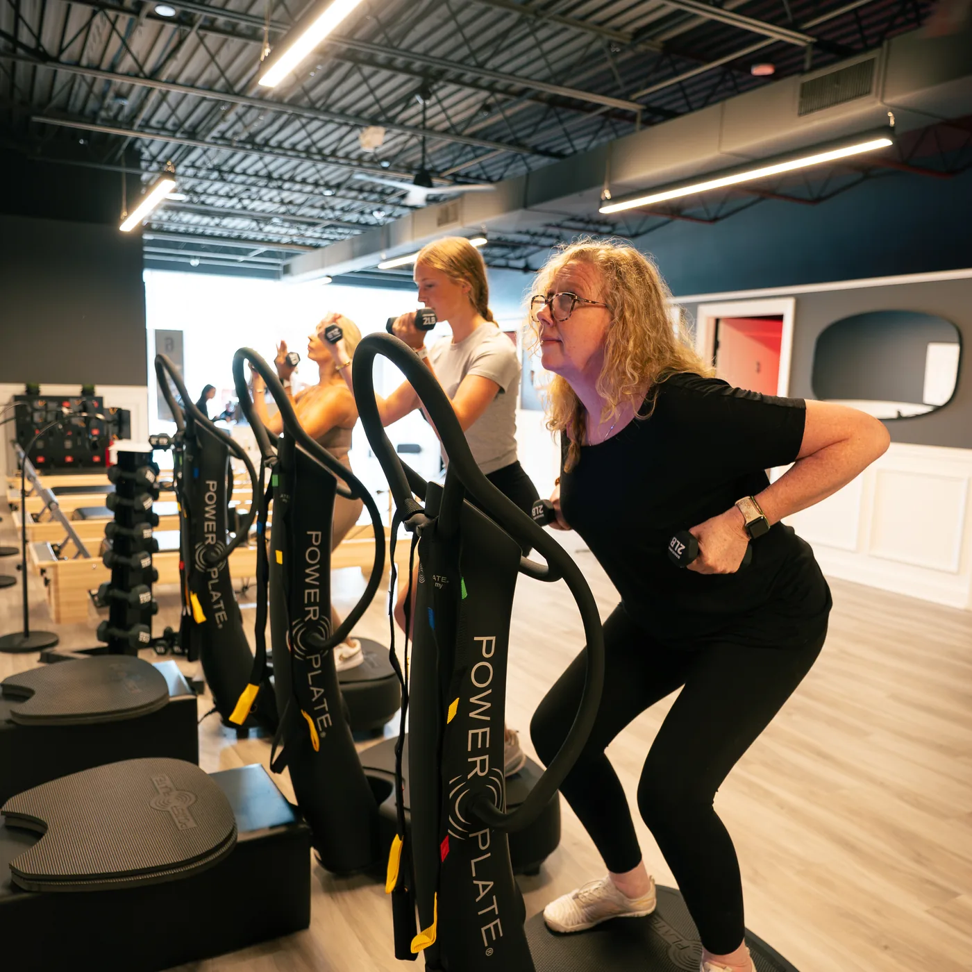 Two women working out on Power Plate whole-body vibration platforms on the open studio floor at Red Light Method Commack