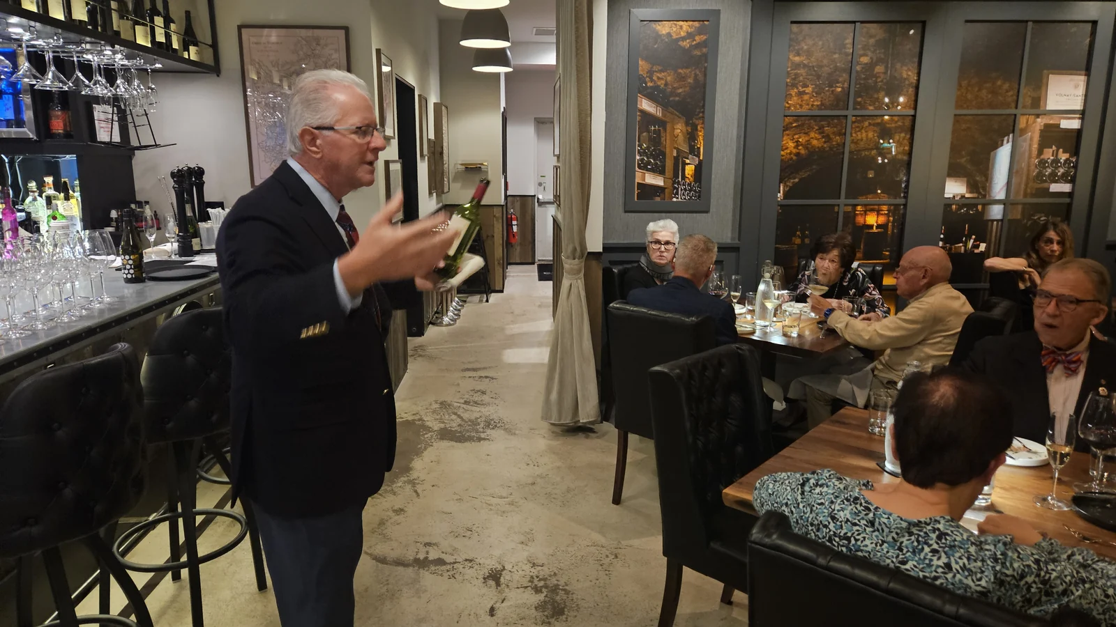 A man in a blazer stands in the dining room at Enology holding a bottle of wine, talking to seated guests at the benefit dinner.