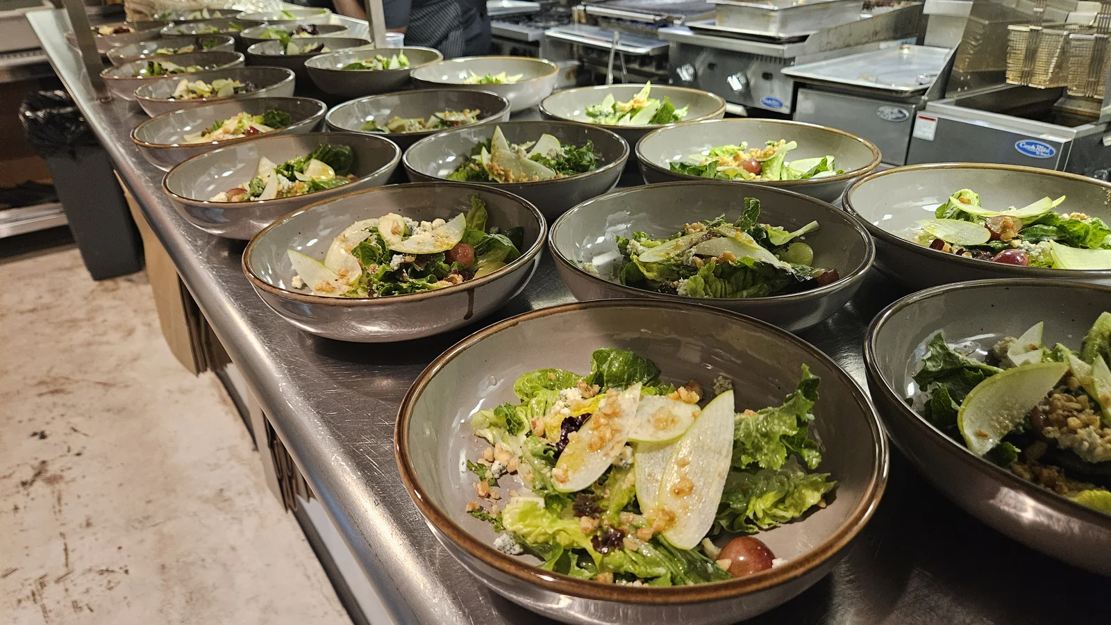 A row of Waldorf salads in shallow bowls lined up along the stainless pass at Enology, apple and pear slices, walnuts, grapes and blue cheese in each one, ready to go out together.