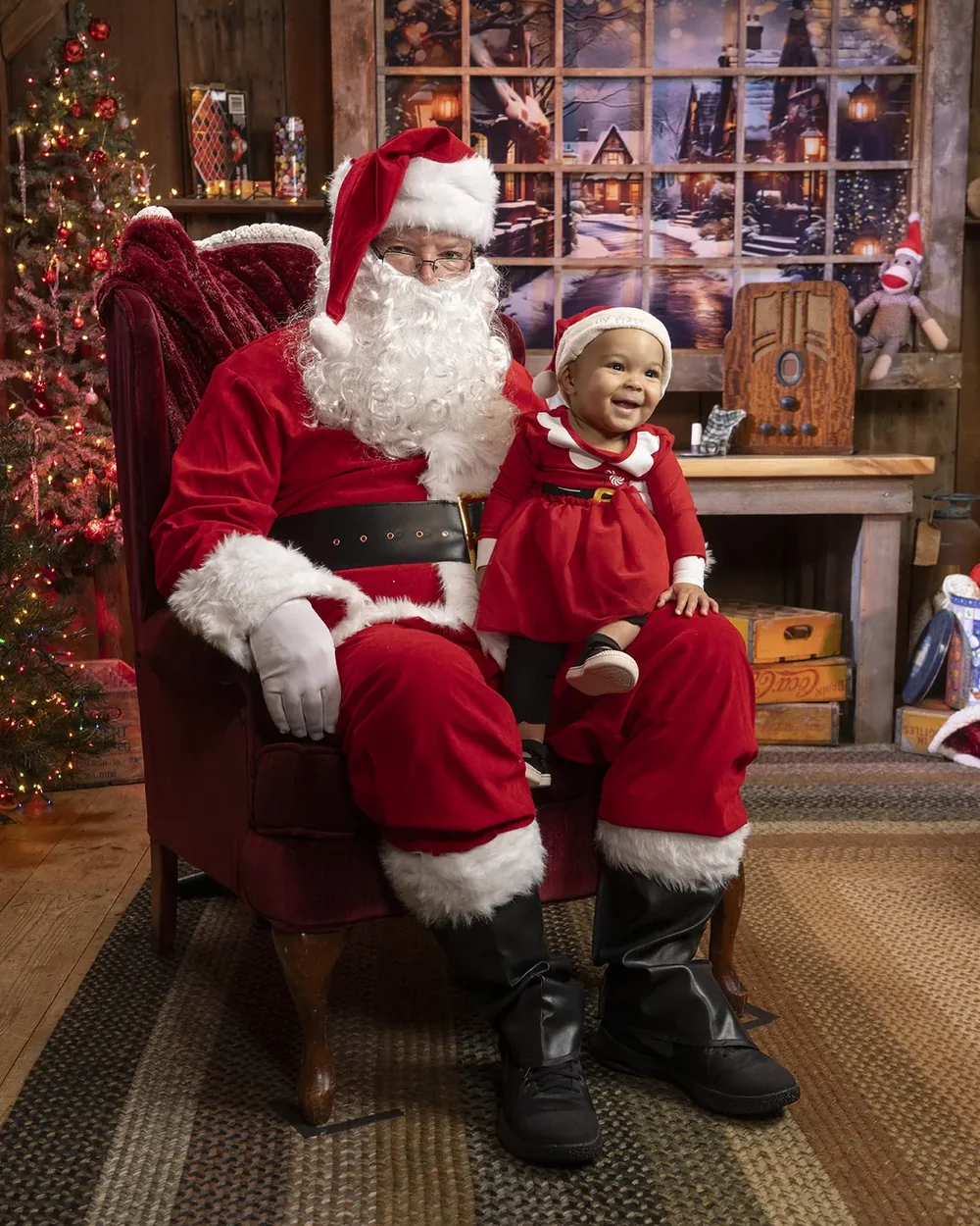 A smiling baby in a red Santa dress sitting on Santa's knee in front of a frosted window and a vintage radio